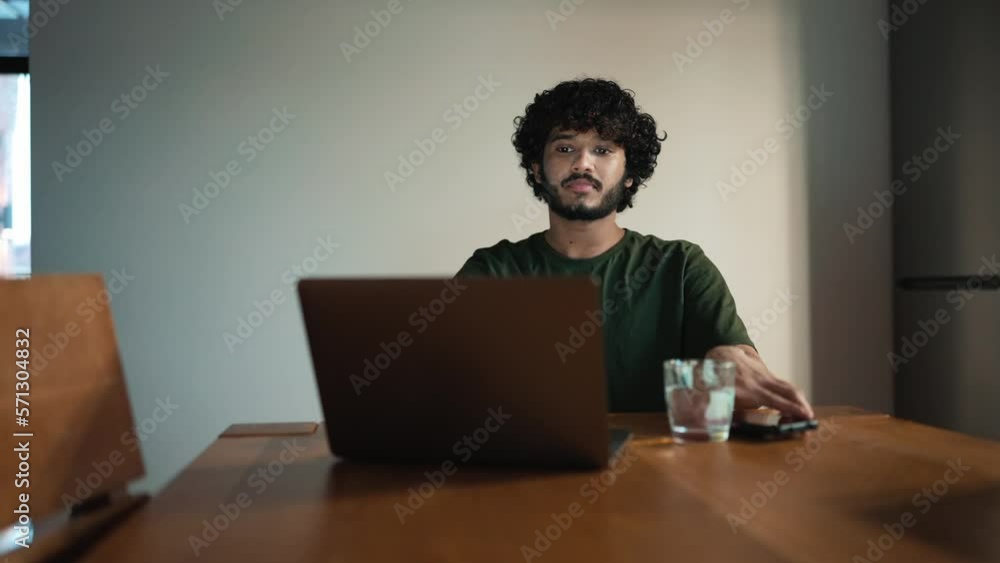 Confident curly haired Indian man working on laptop and looking at mobile in the kitchen at home Confident curly haired Indian man working on laptop and looking at mobile in the kitchen at home