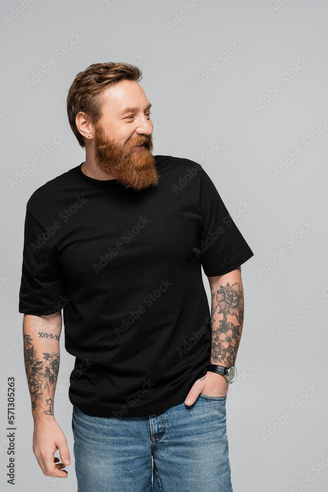 Fototapeta premium cheerful bearded man in black t-shirt standing with hand in pocket of jeans and looking away isolated on grey.