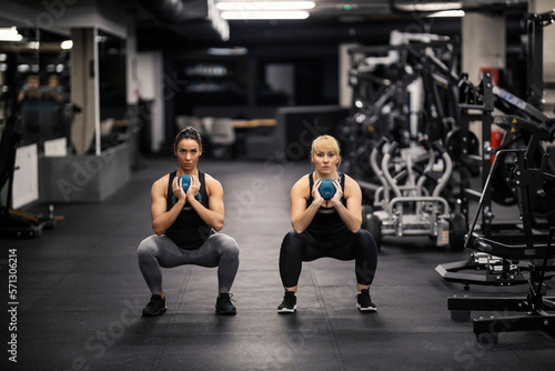 Two strong female bodybuilders are holding kettle bells and doing squats during their strength training in a gym.