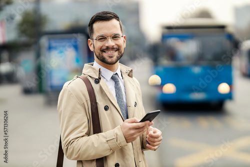 Portrait of a happy elegant man standing on a bus station and waiting for a public bus while using his phone.