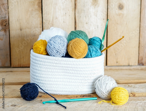 yellow, white and blue woolen balls in a white textile basket with crochet hooks on wooden ground