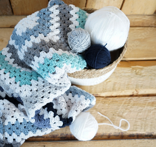 white, grey, blue granny square blanket with woolen balls in an white textile basket on wooden ground