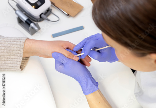 Cropped Nail beautician removes cuticle of female client with orange stick, top view. File, sterilized manicure tools in craft package on white table.Manicure master works in purple gloves.Horizontal