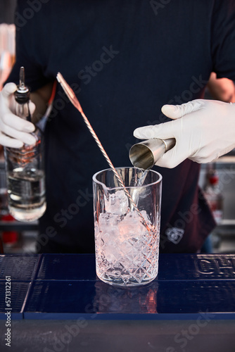 a bartender in white gloves and dark clothes prepares a cocktail, on a bar counter a measuring glass, glass shaker with ice and a bar spoon, a bartender pours alcohol into a measuring glass, jagger photo