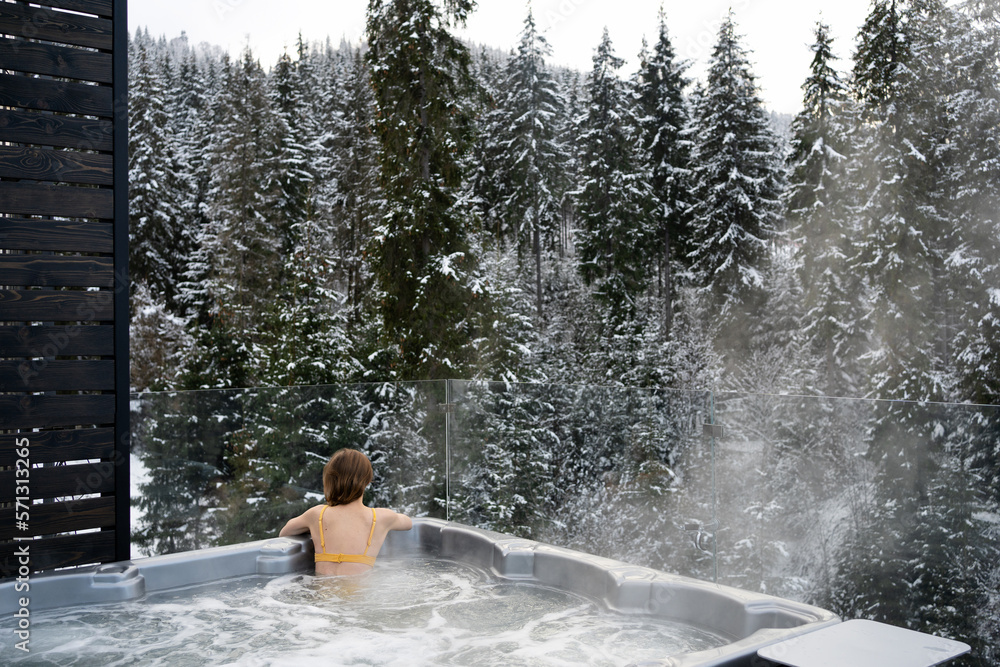 Stockfoto med beskrivningen A young woman relaxes in a hot tub with a  jacuzzi in the winter outdoors, sitting on back and enjoying the view of  the snowy forest. Concept of winter