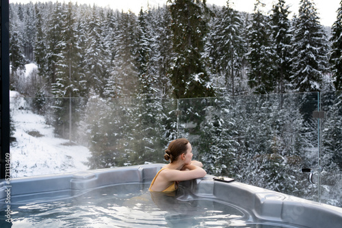 Young woman relaxing in a hot tub and thermal spa enjoying a winter view of snowy Christmas trees. Concept of recreation on hot bath and escape to nature