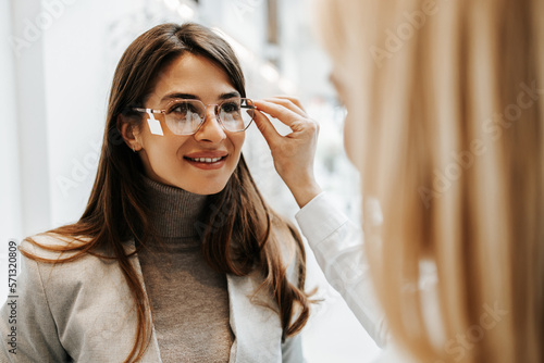 Beautiful and fashionable woman choosing eyeglasses frame in modern optical store. Female seller specialist helps her to make right decision. photo