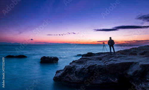 Solo male photographer with camera and tripod watching a blue sunset at sea side on a rock formation