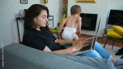 Woman working from home using laptop with child jumping from sofa. Parent in front of computer screen browsing internet online multi tasking