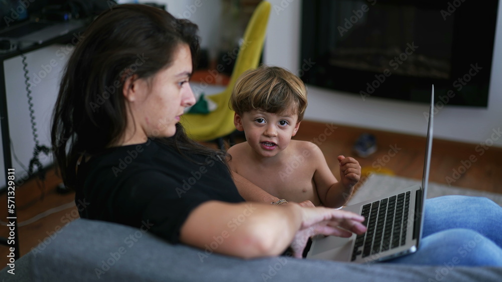Mother parenting with child in front of laptop screen working from home. Parent sitting on couch with toddler boy looking at computer Mother parenting with child in front of laptop screen working from home. Parent sitting on couch with toddler boy looking at computer