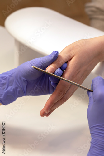 Manicurist shapes, files beautiful woman's nails, holding finger. Caucasian manicure master works in gloves at white work station in spa salon. Soft, safe beauty nail care, treatment. Vertical plane photo