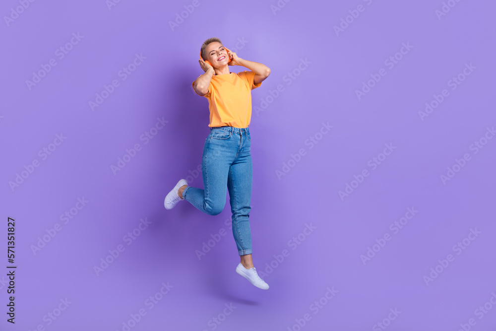Full size photo of optimistic satisfied girl wear sneakers denim pants hands touching headphones isolated on purple color background Full size photo of optimistic satisfied girl wear sneakers denim pants hands touching headphones isolated on purple color background