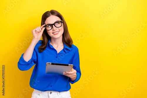 Photo of young lady wear blue shirt brunette hair touch eyeglasses hold tablet look empty space interested offer isolated on yellow color background