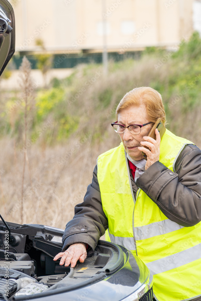 An elderly woman in a reflective vest talks on the phone with the roadside assistance service, next to a damaged car. An elderly woman in a reflective vest talks on the phone with the roadside assistance service, next to a damaged car.