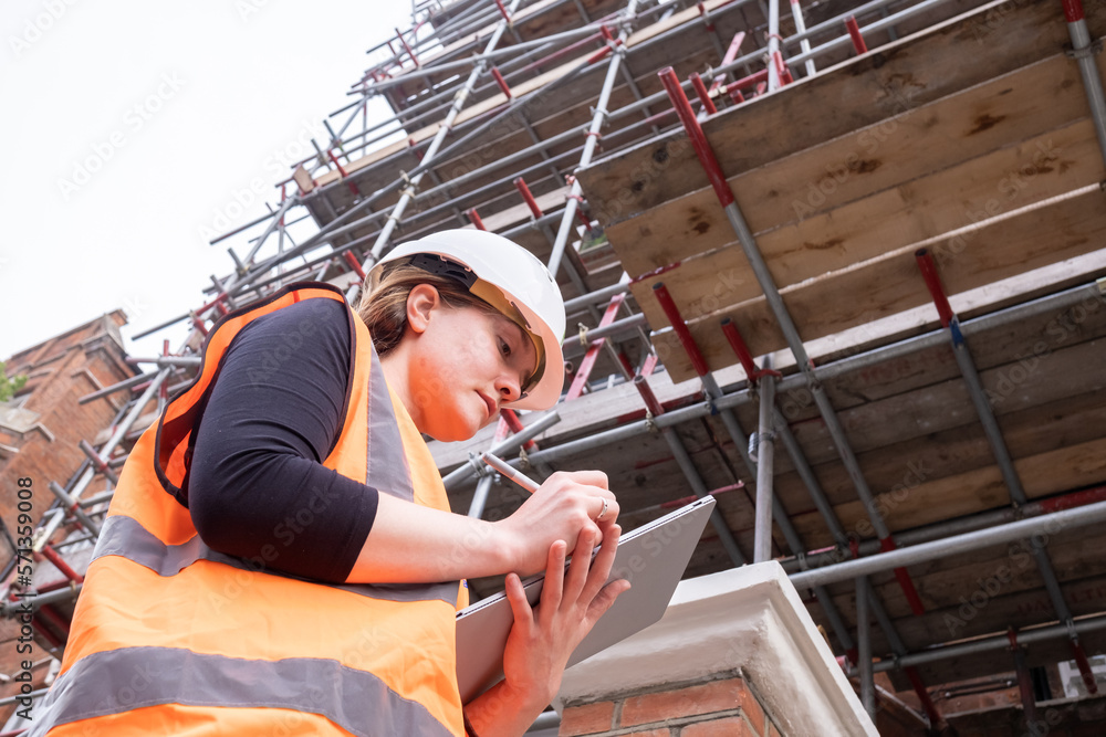 Civil engineer woman writing on a tablet with an electronic pen, hard hat and orange high visibility vest, inspection, touchscreen, technology and innovation in construction site, low angle view