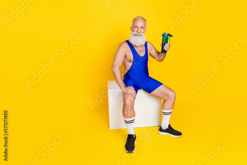 Full length photo of positive sportive grandfather sit platform hold water bottle isolated on yellow color background
