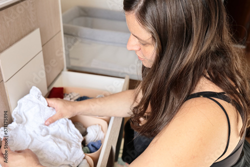 Young pregnant mother lovingly arranges her baby clothes in the drawer chest