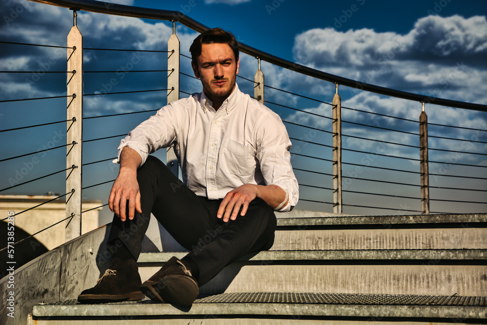 Handsome trendy man wearing white shirt sitting outdoor on metal stairs step Handsome trendy man wearing white shirt sitting outdoor on metal stairs step