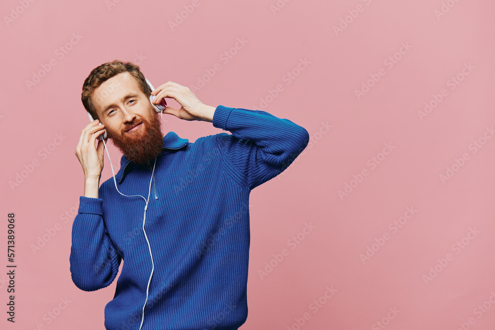 Portrait of a redheaded man wearing headphones smiling and dancing, listening to music on a pink background. A hipster with a beard. Portrait of a redheaded man wearing headphones smiling and dancing, listening to music on a pink background. A hipster with a beard.