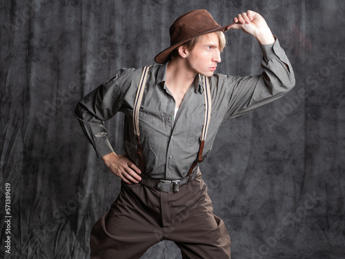 A young man in retro style, an adventure character. A guy in a hat and a gray shirt, breeches with suspenders. photo