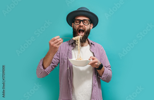 Portrait of a funny cute hipster guy with a beard in a hat opening a big mouth and eating noodles with a fork in the studio.