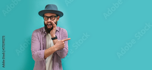 Bilde på lerret A handsome man with a beard in casual clothes wearing a hat and glasses holds his hands near his mouth and tells secret gossip, whispers a malicious conversation, isolated on a blue background