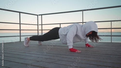 Athlete female doing push up exercises outdoor dressed hoodie sportswear. European athletic female practicing workout on sea embankment. Slow motion full length. Fitness motivation, gym outdoor