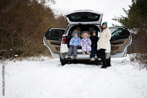 Mother with three children sit on car suv with open trunk stand in winter forest.