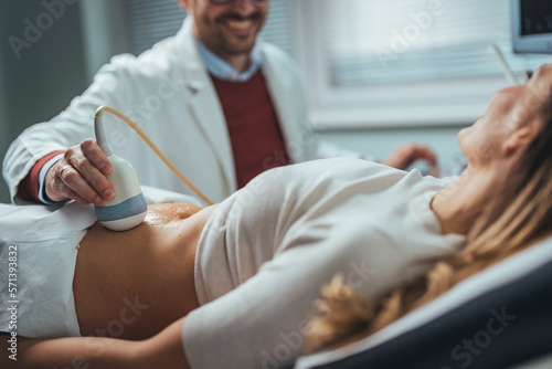 Doctor using ultrasound and screening woman's stomach. Pregnant woman getting ultrasound from doctor. Side view portraits of gynecologist in white lab coat using ultrasound scanner  photo