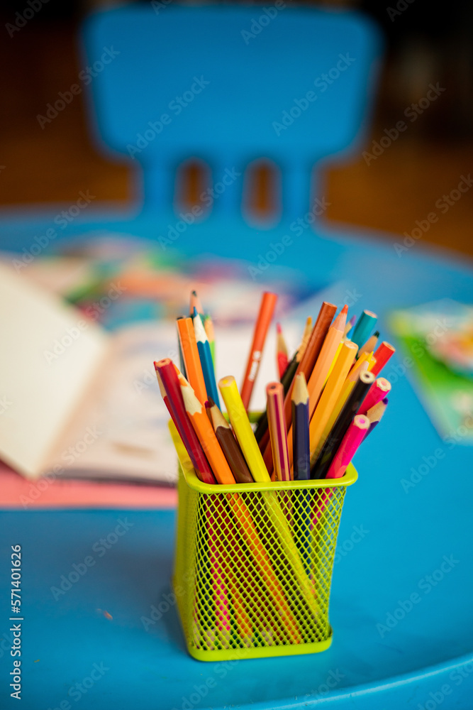 A yellow container with many colored pencils is placed on a blue children's table, next to some coloring books, at a baptism party