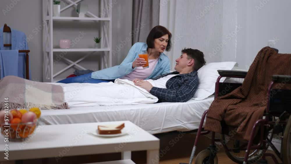 Sick smiling man lying in bed talking to woman entering with orange juice. Loving Caucasian wife helping ill husband drinking refreshing drink in bedroom with wheelchair Sick smiling man lying in bed talking to woman entering with orange juice. Loving Caucasian wife helping ill husband drinking refreshing drink in bedroom with wheelchair