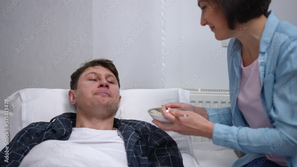 Zoom in to happy Caucasian husband lying in bed as loving wife feeding spouse with milk porridge. Smiling grateful man in bedroom at home with caring woman. Illness and bonding concept Zoom in to happy Caucasian husband lying in bed as loving wife feeding spouse with milk porridge. Smiling grateful man in bedroom at home with caring woman. Illness and bonding concept