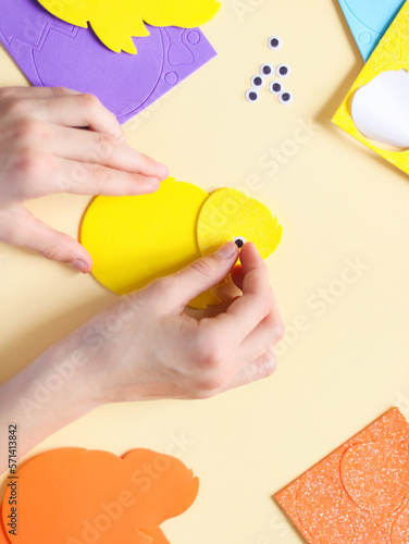 The hand of a caucasian teenage girl glues a decorative eye on a felt yellow chick