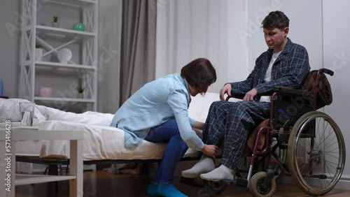 Wide shot loving wife helping husband in wheelchair putting feet on footrest gesturing high-five. Positive Caucasian man and woman together at home indoors