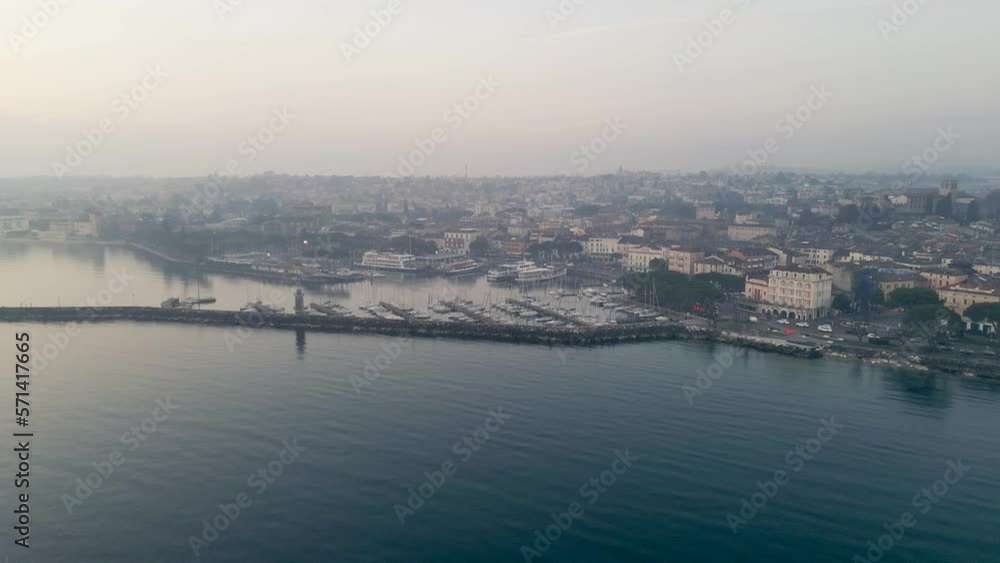 drone flight garda lake near town desenzano del garda in the morning, fog hangs over lake and harbor with boats, beautiful vacation destination in italy, road traffic rushes by. drone flight garda lake near town desenzano del garda in the morning, fog hangs over lake and harbor with boats, beautiful vacation destination in italy, road traffic rushes by.