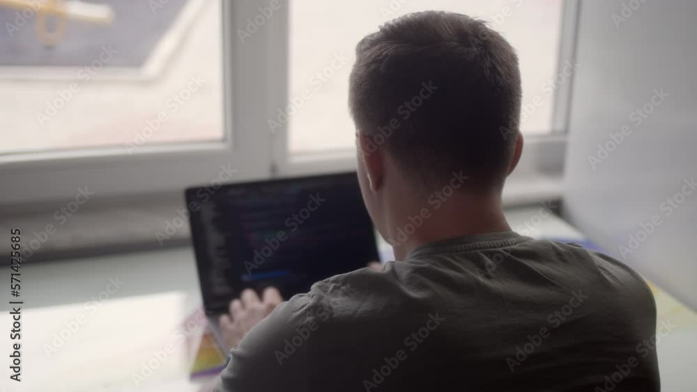 Rear view of man who works on laptop, programmer. IT specialist at home office work in front of bright light window. professional web designer programs back end site. Rear view of man who works on laptop, programmer. IT specialist at home office work in front of bright light window. professional web designer programs back end site.