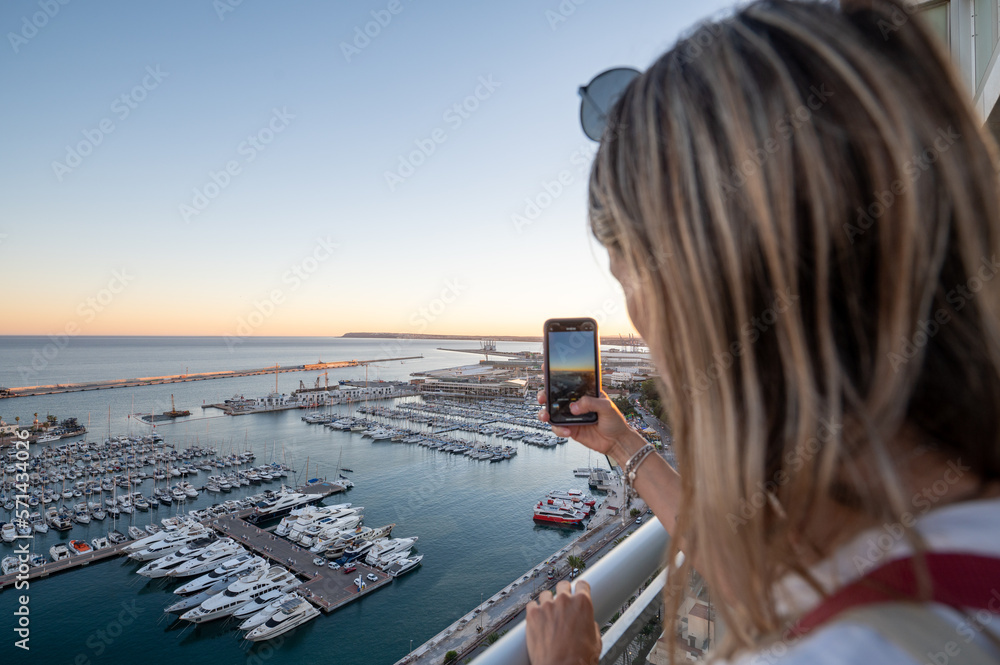 Woman takes a photo of the Port of Alicante from a hotel terrace in Spain.