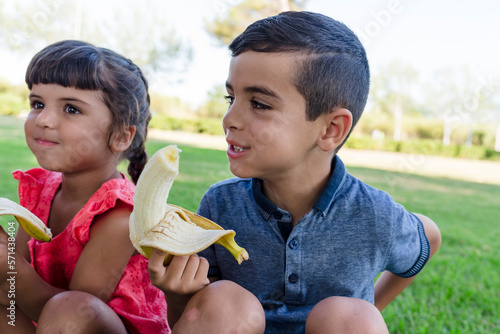 Two kids eating banana sitting outdoors on the grass in a park.