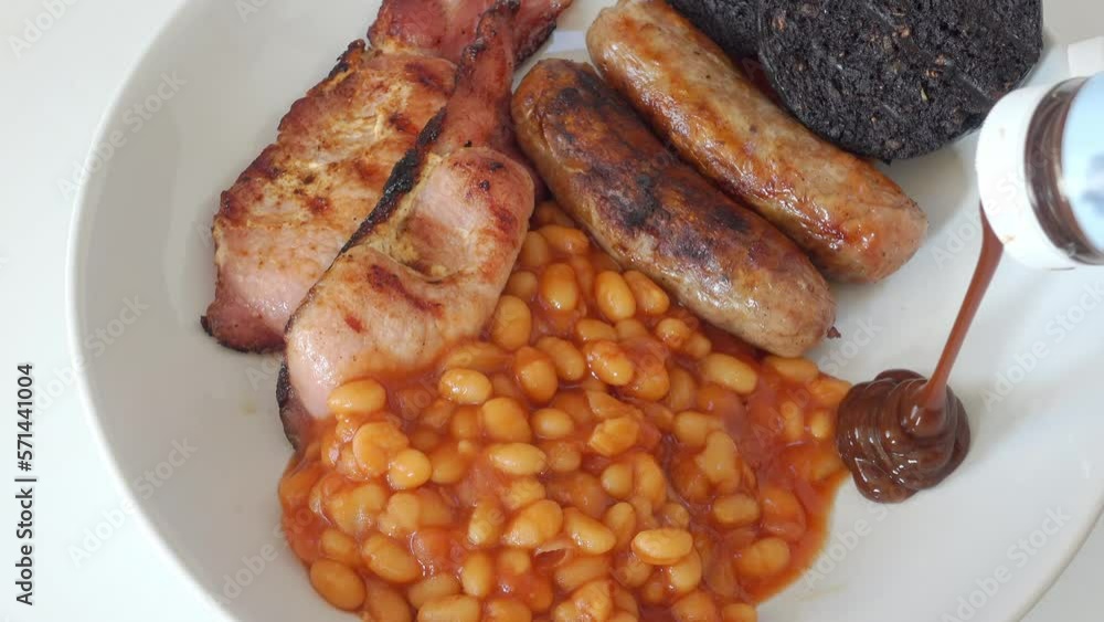 Brown sauce being added to a traditional English breakfast of cumberland sausages, back bacon black pudding and baked beans, but no egg, on a white plate. Real time, zoom out. Brown sauce being added to a traditional English breakfast of cumberland sausages, back bacon black pudding and baked beans, but no egg, on a white plate. Real time, zoom out.