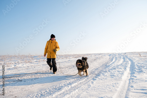 Male in yellow coat walking with his big black dog on winter background. Family winter activity with pet on sunny day outdoor. Mongolian dog breed. 