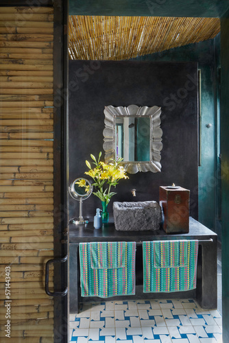 Baño rústico de paredes y techo de carrizo, con mesa y paredes de cemento pulido donde un espejo de latón armoniza con un florero de lilis amarillas y una pileta de piedra volcánica . photo