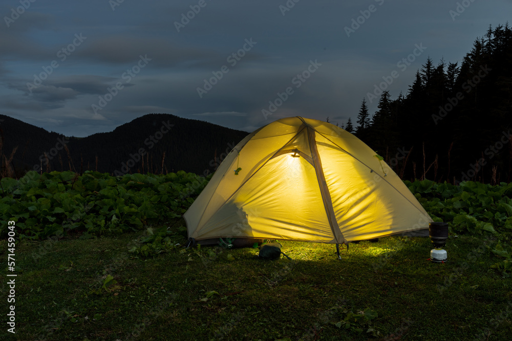 Luminous tourist tent in nature. Tourist tent and equipment. Camping. Tent at dusk. Glowing tent against the backdrop of mountains.