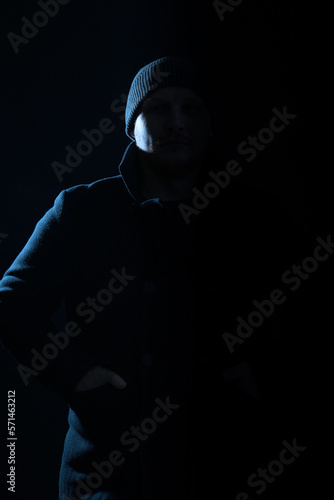Horror concept. Studio portrait of man with dark coat and hat hiding in black shadow. Model silhouette illuminate with light. Copy space and dark studio background. Toned image with blue color