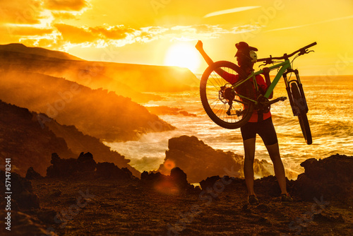 Success, achievement, accomplishment and winning concept with cyclist mountain biking. Happy MTB woman cycling raising arms lifting bike by sea during sunset cheering and celebrating at summit top.