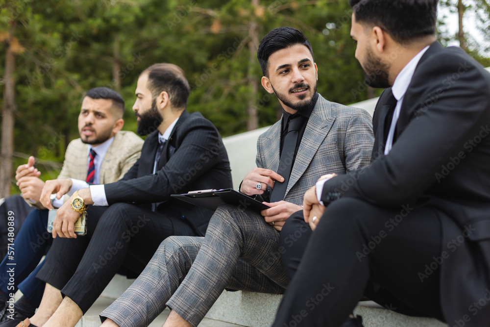 Business people outdoor meeting. Several men in suits sit on the steps of a street staircase during a work break. Successful promotion and recognition in the team of employees. Business people outdoor meeting. Several men in suits sit on the steps of a street staircase during a work break. Successful promotion and recognition in the team of employees.