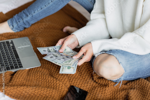 Young caucasian woman holding hands cash money dollars bills. Person counting money at home on bed, next lying laptop and mobile phone.
