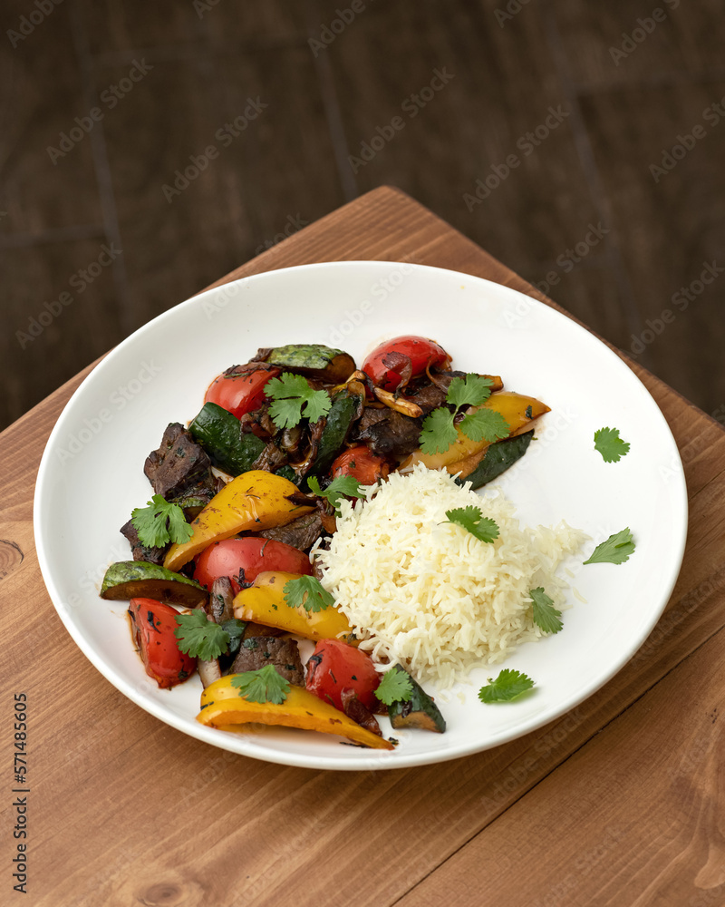 Baked meat with vegetables and boiled rice for garnish. Dish is decorated with parsley in white plate on wooden table. Complex lunch. View from above. Copy space. Soft focus. Baked meat with vegetables and boiled rice for garnish. Dish is decorated with parsley in white plate on wooden table. Complex lunch. View from above. Copy space. Soft focus.