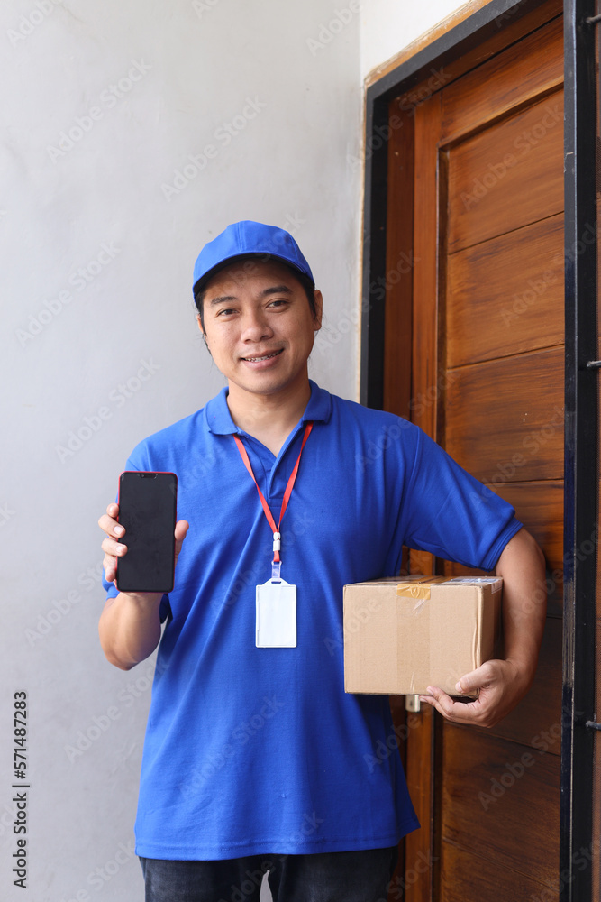 Smiling young deliveryman in blue uniform deliver box in front of the door while showing blank screen of smartphone for mock up and copy space. Smiling young deliveryman in blue uniform deliver box in front of the door while showing blank screen of smartphone for mock up and copy space.