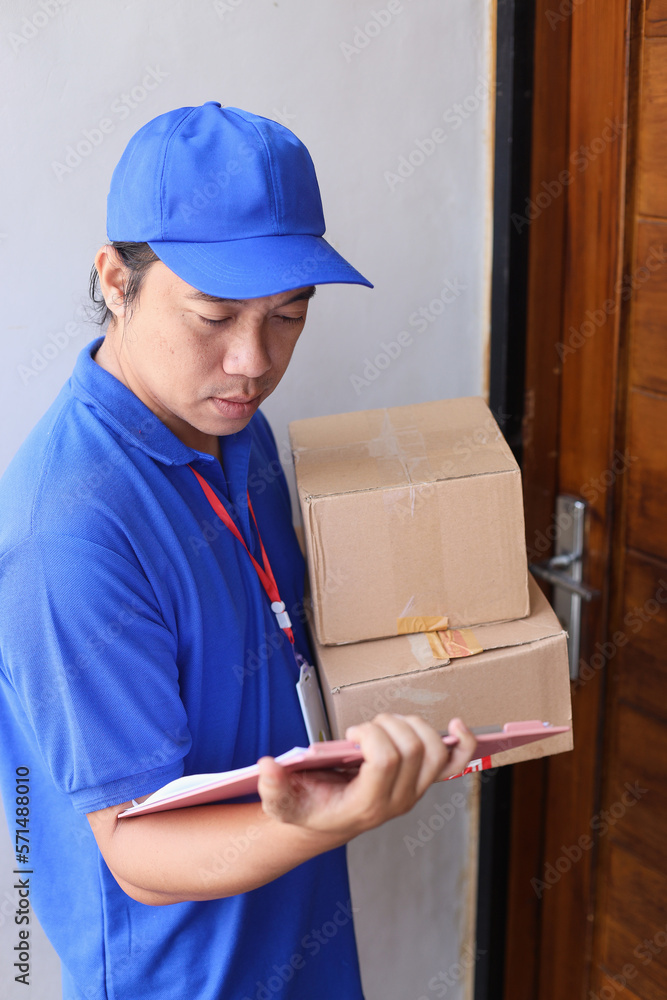 Top view of deliveryman in blue uniform wearing cap is holding pile of cardboard and checking document in front of the door, sending package to customer. Top view of deliveryman in blue uniform wearing cap is holding pile of cardboard and checking document in front of the door, sending package to customer.