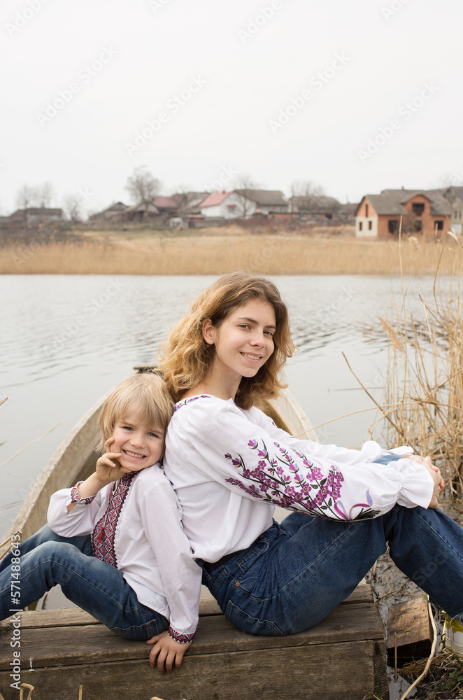Fototapeta premium happy little boy and young beautiful woman in embroidered national clothes sit next to each other on shore of lake. Family, refugees, unity, support, freedom, independence. Ukrainians against the war